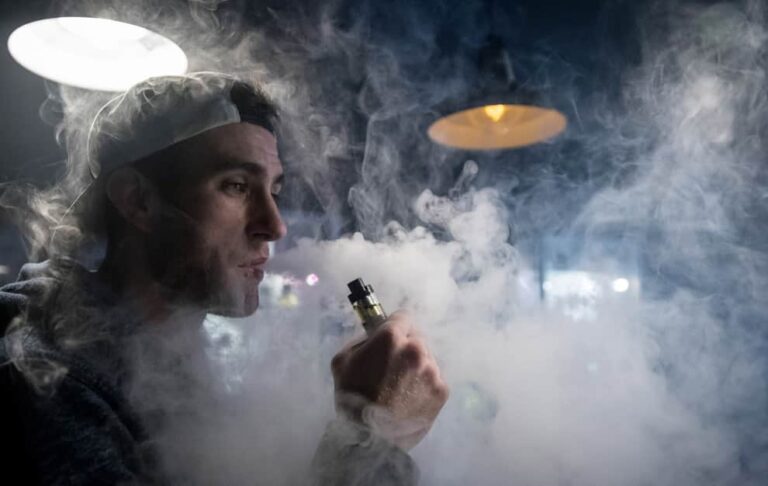 A man in a backward white cap exhales dense clouds of vapor while using a vape device in a dimly lit room with industrial-style hanging lights.